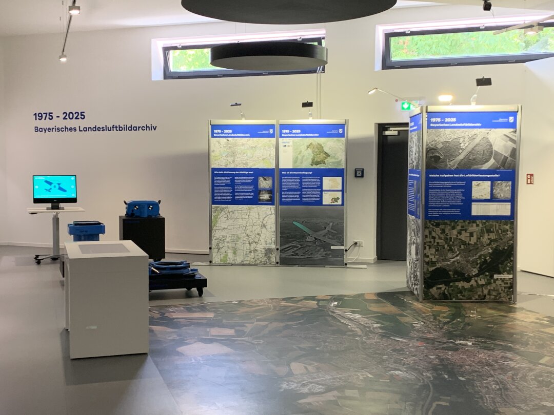 In der Bildmitte und rechts stehen Wall Ups, die Luftbilder, Karten und ein Flugzeug zeigen. Links steht die Recherchestation und dahinter der Erklärfilm zu Luftbildfotografie. Links, mittig stehen noch die RMA-Kamera in  drei einzelnen Teilen.